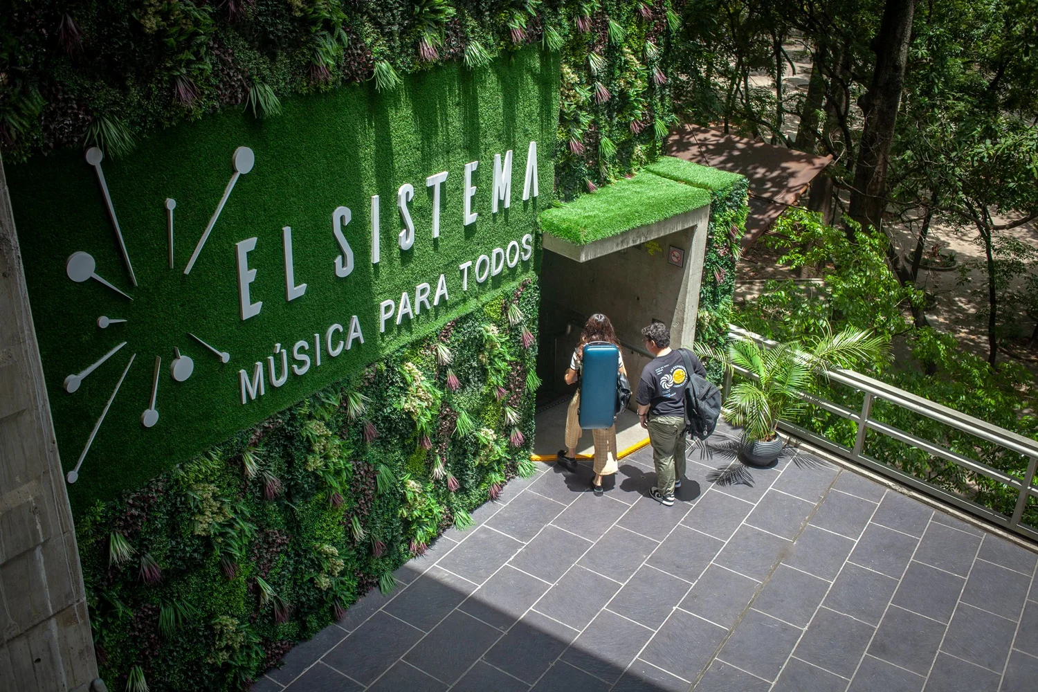 Un muro verde con el cartel «El Sistema, música para todos» y dos jóvenes que entran